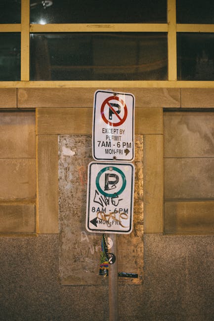 A tall metal parking restriction sign mounted on a pole, positioned against a building wall made of beige stone with a large window above. The top sign indicates no parking except by permit from 7 am